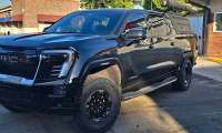 Black GMC Sierra Denali with off-road tires and an aftermarket bed cap parked outside a storefront.