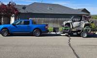 Blue Ford Maverick towing a side-by-side off-road vehicle on a trailer in a residential street.
