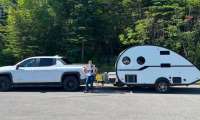 White Chevrolet Silverado EV towing a small white teardrop camper trailer, parked at a forested campsite