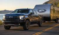 Black Chevrolet Silverado pickup towing a Grey Wolf travel trailer on a road beside a lake and mountains