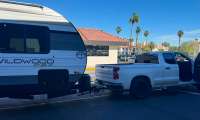 White Chevrolet Silverado pickup with door open, towing a Wildwood FSX travel trailer in a parking lot with palm trees
