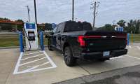 Black Ford F-150 Lightning charging at a GM Energy DC fast charger near a gas station.