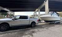 Ford F-150 Lightning towing a large cabin boat on a trailer under a bridge near the water.