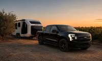 Black Ford F-150 Lightning towing a white Pebble Flow travel trailer through a vineyard at sunset.
