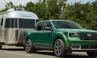 Green Ford Maverick pickup truck towing a silver Airstream travel trailer on a tree-lined highway