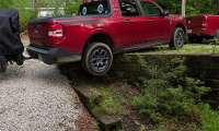 Red Ford Maverick Hybrid XLT towing a covered motorcycle trailer, rear view driving over a low stone retaining wall