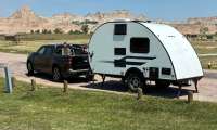 Black Ford Maverick towing a white teardrop camper with Badlands rock formations in the background.