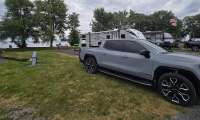 Silver GMC Sierra EV Denali pickup truck parked at a lakeside campground, with a Bullet travel trailer and lake visible in the background