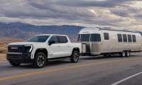 White GMC Sierra EV towing an Airstream travel trailer on a desert highway with mountains in the background.