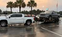 White GMC Sierra pickup towing another GMC Sierra on a trailer in a wet dealership parking lot.