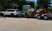 White Kia Telluride SUV towing a flatbed trailer loaded with a compact tractor on a suburban street