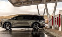 Lucid Gravity electric SUV charging at a Tesla Supercharger station, side profile with multiple charging stalls in a desert landscape.