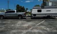 Silver Ram 1500 pickup towing a small Coleman travel trailer in a parking lot with palm trees