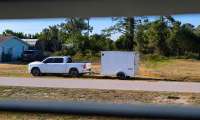 White Ram 1500 pickup towing a small enclosed cargo trailer, parked on a roadside near trees and a house