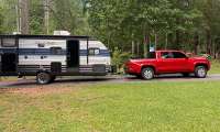 Red Toyota Tacoma towing a Wolf Pup travel trailer on a wooded driveway near a campsite.