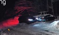 Ring camera footage at night showing a dark Ford Mustang Mach-E with bright headlights and red taillights, and a dark sedan, stuck or maneuvering in a snow-covered driveway.