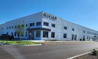 Rivian facility exterior with a large Rivian sign on the building, parked vehicles, and an empty parking lot under a clear blue sky.