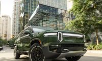Rivian R1S electric SUV driving through a modern city street with glass skyscrapers and urban traffic.