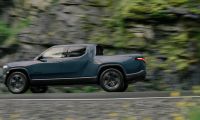 Side view of a Rivian R1T electric truck driving on a road with a rocky, green hillside background, motion blur showing speed.