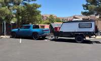 Blue Rivian R1T towing an off-road camper trailer while parked beside EV charging stations.