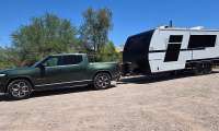 A green Rivian R1T electric pickup towing a white and black Brinkley travel trailer on a gravel lot.