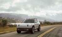 White Rivian R2 driving on a two-lane road with cloudy hills in the background.