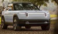 Silver Rivian R2S parked on a wooded trail in a front three-quarter view.