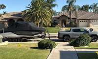 Silver GMC Sierra EV towing a covered boat past large homes and palm trees in a residential neighborhood.