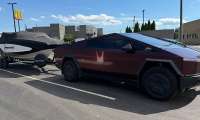 Tesla Cybertruck towing a MasterCraft boat while parked beside Tesla Supercharger stalls in a sunny parking lot.