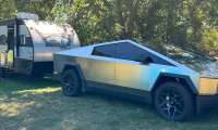 Tesla Cybertruck towing a Patriot Edition travel trailer, parked on grass under shade trees with a dog resting nearby