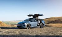 Blue Tesla Model X electric SUV with falcon-wing doors open, parked on a scenic overlook in a rural landscape.