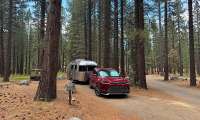 Red Toyota Grand Highlander towing an Airstream travel trailer through a pine forest campsite.