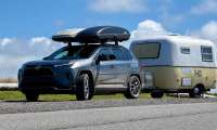 Toyota RAV4 towing a small vintage travel trailer while carrying a large rooftop cargo box under a blue sky.