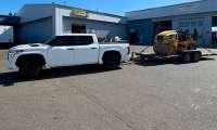White Toyota Tundra pickup towing a flatbed trailer loaded with a CAT compactor roller outside an industrial equipment yard