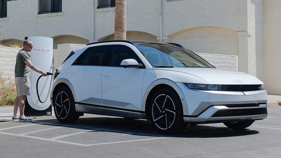 2024 Hyundai Ioniq 5 in white parked at a Tesla Supercharger station with an owner plugging in the NACS charging cable demonstrating the Ioniq 5s expanded charging network access in a sunny suburban parking lot