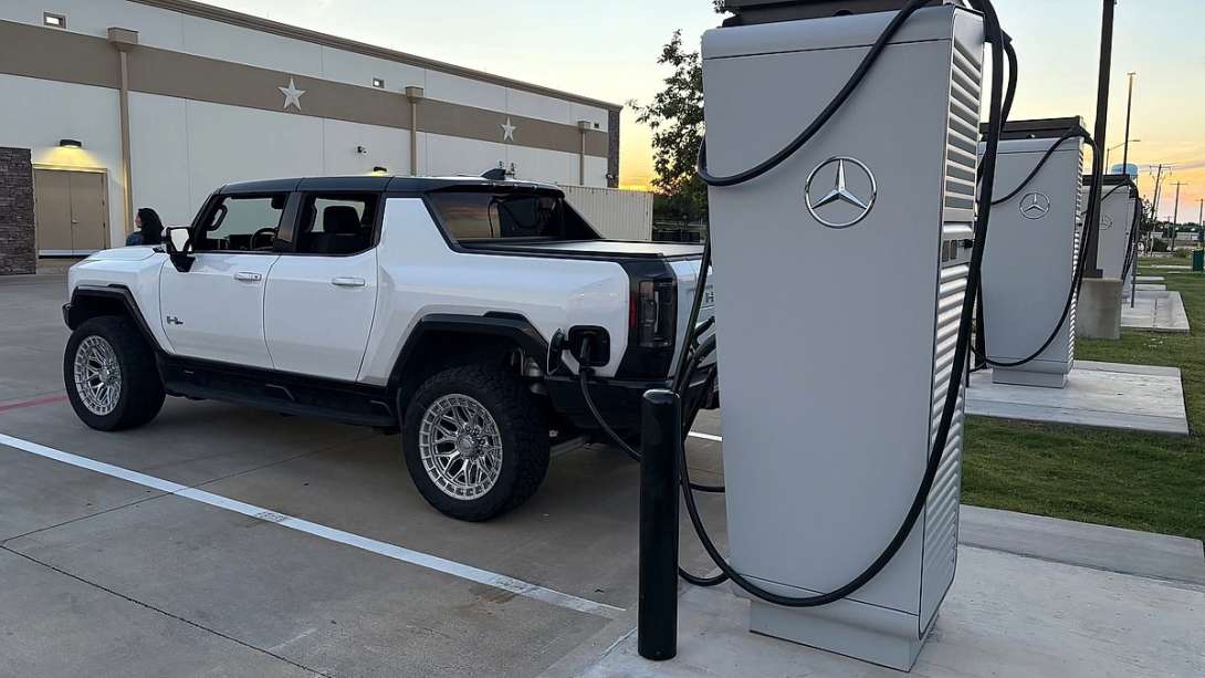 White GMC Hummer EV pickup charging at a Mercedes-Benz charging station at sunset.