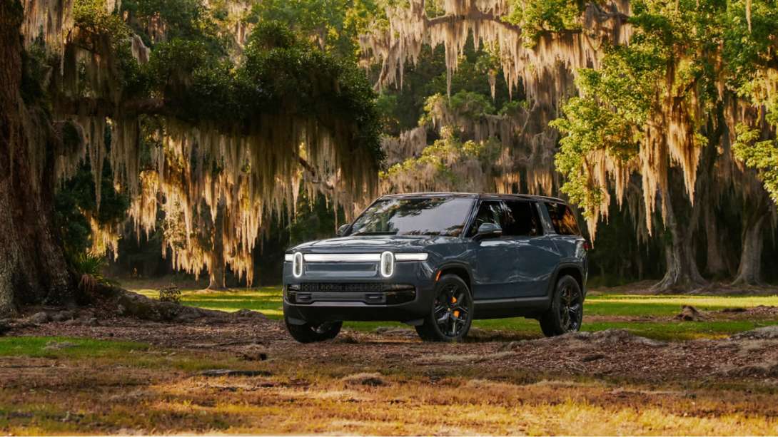 Gray-blue Rivian R1S SUV parked under oak trees draped in Spanish moss, front three-quarter view