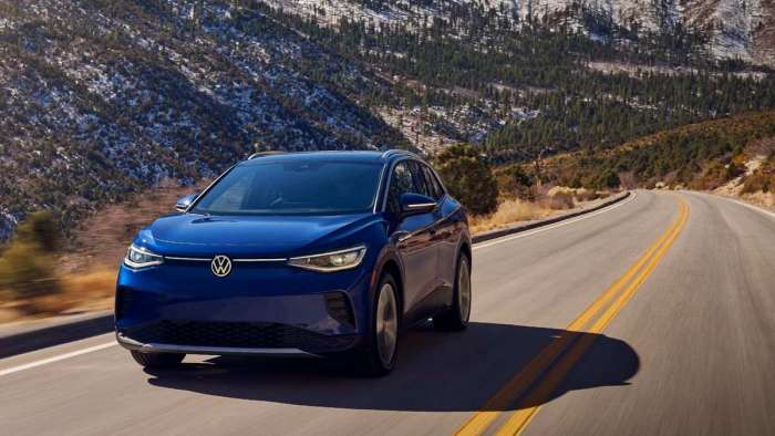 A blue Volkswagen ID.4 is pictured driving on a long, straight road with snow-dusted mountains and trees in the background.